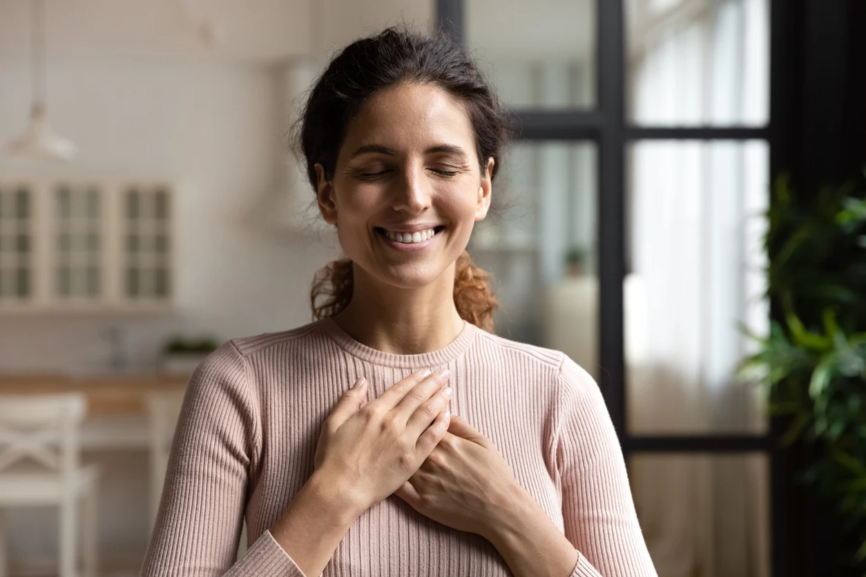 Mulher com os olhos fechados sorrindo com as mãos na altura do coração